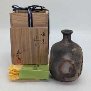 May include: A dark brown ceramic sake bottle with a narrow neck, next to a wooden box with Japanese characters and a blue ribbon. The box is open, revealing a green paper and yellow cloth.