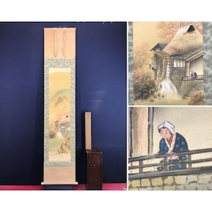 Op de afbeelding: Een Japanse rolschildering die een landelijk landschap met een traditioneel huis en bergen afbeeldt. Het kunstwerk heeft zachte kleuren en een serene sfeer. De rol wordt getoond met een houten kist en een donkerblauwe achtergrond.