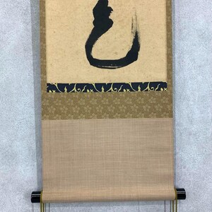 May include: A traditional Japanese scroll painting with a large black calligraphic character on a tan background. The scroll features decorative borders in dark blue and gold, and a tan fabric bottom.