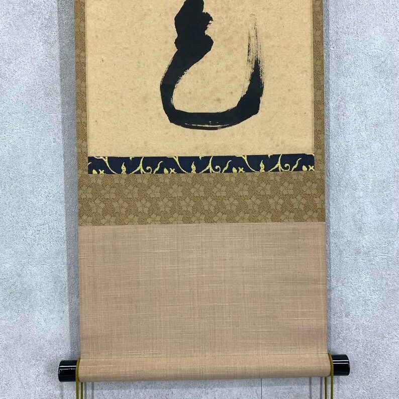 May include: A traditional Japanese scroll painting with a large black calligraphic character on a tan background. The scroll features decorative borders in dark blue and gold, and a tan fabric bottom.