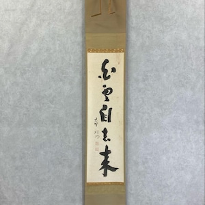 May include: A vertical scroll featuring Japanese calligraphy in black ink on a cream background. The scroll is framed with gold trim and has a tan backing. It has a hanging loop at the top and a wooden bar at the bottom.