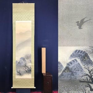Op de afbeelding: Een Japanse rolschildering met een winterlandschap met bergen, bomen en een vogel in vlucht. De rol heeft een lichtgroene rand met een bloemmotief aan de bovenkant. Een houten blok en een donkerbruine doos staan ernaast.