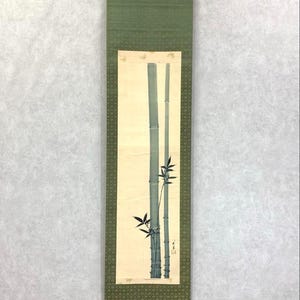 May include: A vertical Japanese scroll painting featuring two bamboo stalks in shades of blue-green against a cream background. The scroll is framed by a green patterned border and a solid green top section. The artwork is suspended by a metal rod.