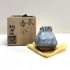 May include: A ceramic incense holder shaped like a small house with a light blue glaze. It sits on a yellow cloth next to a wooden box with Japanese characters and a seal. The holder has a rustic, handmade appearance.