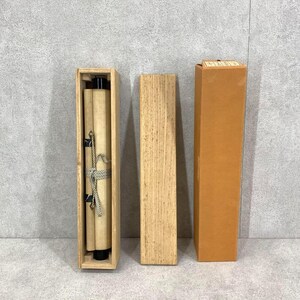 May include: A traditional Japanese scroll painting in a wooden box. The scroll is rolled up and tied with a decorative cord. The box is rectangular and made of light-colored wood. A separate wooden lid and a brown cardboard box are also present.