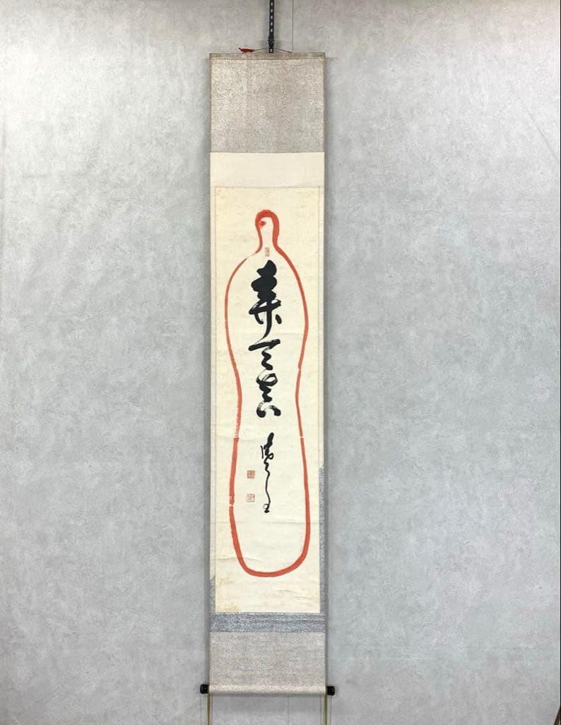 May include: A vertical Japanese scroll with calligraphy in black ink and a red outline. The scroll features a cream-colored background with a red outline in the shape of a bottle. The top and bottom sections have a silver-gray textured design.