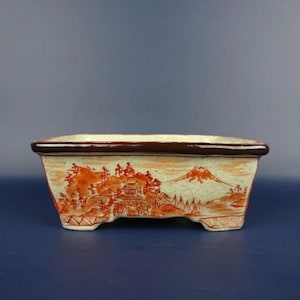 May include: Rectangular ceramic bonsai pot with a brown rim and feet. The pot features a hand-painted landscape scene in shades of orange and brown, depicting mountains, buildings, and trees against a light green background.
