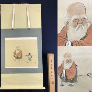 Op de afbeelding: Een antiek Japans rolschilderij met een oude man met een lange witte baard en een jonge figuur. Het kunstwerk is in gedempte tinten oranje, groen en beige, met een houten opbergdoos. De rol heeft witte linten om op te hangen.