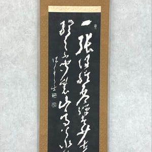 May include: A traditional Japanese scroll featuring white calligraphy on a black panel, set within a brown border. The scroll is finished with a wooden rod at the base. The text is arranged vertically.