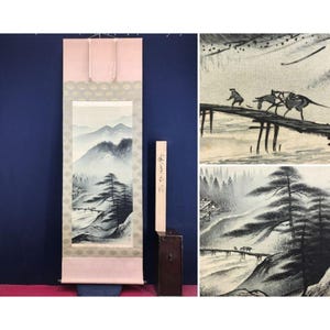 May include: A Japanese scroll painting depicting a misty mountain landscape with a bridge and figures. The artwork is in shades of gray and black, with a pink and gold border. A wooden box and a wooden stick with Japanese characters are included.