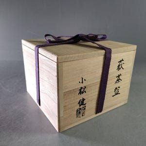 May include: A light-colored wooden box with a purple ribbon tied around it. The box has Japanese characters printed on the side. The box is square and has a lid. The ribbon is tied in a bow on top.