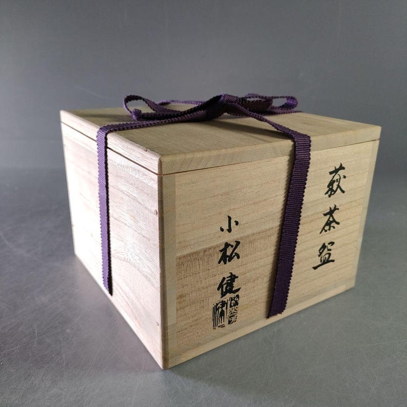 May include: A light-colored wooden box with a purple ribbon tied around it. The box has Japanese characters printed on the side. The box is square and has a lid. The ribbon is tied in a bow on top.