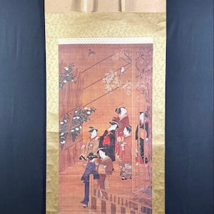 Op de afbeelding: Een verticale hangende rol met een gedetailleerde schildering van verschillende figuren in traditionele kleding op een houten veranda. Het kunstwerk is omlijst door goudkleurige stoffen randen met een subtiel patroon en heeft twee ophangbanden.