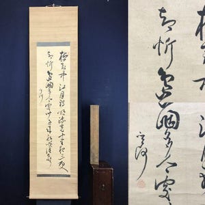 Op de afbeelding: Een traditionele Japanse rol met kalligrafie in zwarte inkt op crèmekleurig papier. De rol hangt tegen een donkerblauwe achtergrond. Een houten kist en een houten stok staan naast de rol.