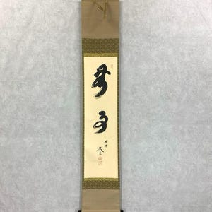 May include: A vertical Japanese scroll with black calligraphy on a cream-colored background. The scroll is framed by decorative borders and a tan backing. The calligraphy features two bold characters, with additional smaller text below.
