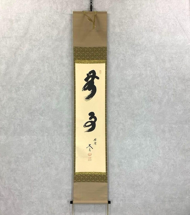 May include: A vertical Japanese scroll with black calligraphy on a cream-colored background. The scroll is framed by decorative borders and a tan backing. The calligraphy features two bold characters, with additional smaller text below.
