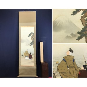 May include: An antique Japanese scroll painting depicting a landscape with Mount Fuji and figures in traditional dress. The artwork is displayed vertically, with a neutral border and wooden elements. The painting shows a tranquil scene with soft colours.