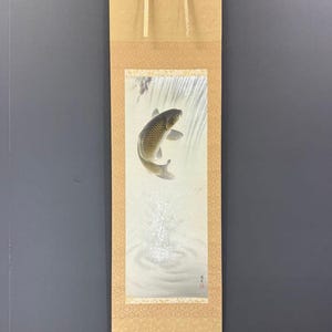 May include: A vertical hanging scroll featuring a painting of a grey koi fish leaping from water. The fish is depicted mid-air with detailed scales, while ripples and splashes are visible below. The artwork is mounted on a light tan fabric border.