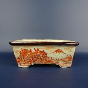 以下が含まれることがあります： 茶色の縁と足が付いた長方形のセラミック盆栽鉢。オレンジと赤の色合いで手描きされた風景画が特徴で、山、建物、木々が淡い緑色の背景に描かれています。