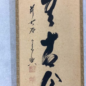 May include: A vertical scroll featuring Japanese calligraphy in black ink on a beige background. The calligraphy includes several characters and a red stamp. The scroll is framed with a brown border.