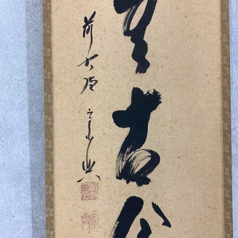 May include: A vertical scroll featuring Japanese calligraphy in black ink on a beige background. The calligraphy includes several characters and a red stamp. The scroll is framed with a brown border.