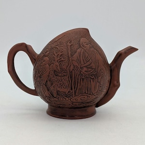 以下が含まれることがあります： 茶色のセラミックティーポット。ひげを生やした人物、杖、鹿、別の人物の精巧なレリーフ彫刻が施されています。 ティーポットは、湾曲したハンドルと注ぎ口を備え、白い背景に配置されています。