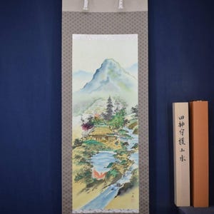 May include: A Japanese scroll painting showing a mountain scene with a house, river, and trees. The artwork uses a soft colour palette with greens, blues, and browns. The scroll has a patterned border and white ribbons for hanging. The painting is approximately 100 cm in length.