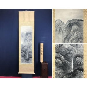 May include: A Japanese scroll painting showing a monochrome mountain scene. The artwork is mounted on a beige background with gold detailing. A wooden box with Japanese text is positioned next to the scroll.