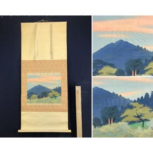 May include: A vertical hanging scroll featuring a landscape painting of a blue mountain range under a soft pink and blue sky, with green trees in the foreground. The scroll has a cream-colored border and includes a matching storage box.