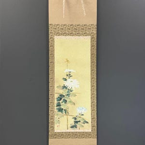 May include: A vertical hanging scroll featuring a painting of white chrysanthemum flowers and a small dragonfly on a pale yellow background. The artwork is framed by a patterned fabric border in shades of brown and gold, with light tan fabric at the top and bottom ends.