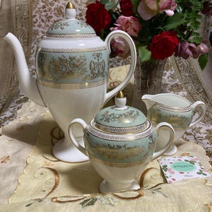 Könnte beinhalten: Ein Vintage-Teeservice mit Teekanne, Zuckerdose und Sahnekännchen. Das Set hat ein weißes und hellgrünes Farbschema mit goldenen floralen Akzenten. Die Teekanne hat ein hohes, schlankes Design.