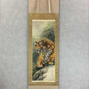 May include: A traditional Japanese scroll painting featuring a tiger. The artwork depicts a detailed tiger with orange and black stripes, perched on a rock formation. The scroll has a decorative border and is ready to hang.