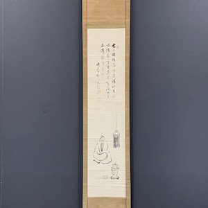 May include: A vertical hanging scroll featuring a light beige paper panel with black ink brush paintings of a seated figure, a hanging lantern, and a tea set. The upper section contains vertical columns of black calligraphy and red seal stamps.