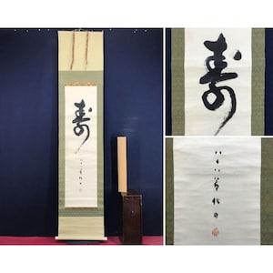 Op de afbeelding: Een lange, verticale Japanse rol met zwarte kalligrafie op wit papier. De rol heeft een crèmekleurige boven- en onderkant met een groen patroon. Een kleine houten kist staat aan de zijkant.