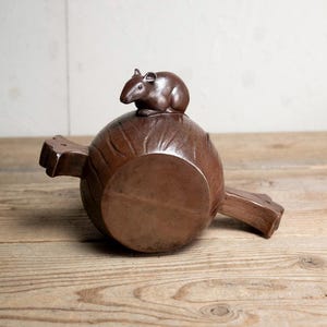 Könnte beinhalten: Ein brauner, dekorativer Nussknacker mit rundem Sockel und zwei Griffen. Eine kleine, braune Mäusefigur sitzt auf dem runden Sockel. Der Nussknacker ist aus Holz gefertigt und hat ein rustikales Aussehen.