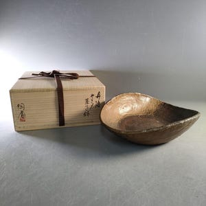 May include: A handmade, brown ceramic bowl with a unique, organic shape. The bowl has a textured surface and a dark brown interior. Beside it is a wooden box tied with a brown ribbon, with Japanese text.