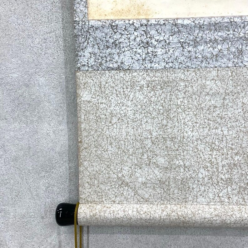 May include: A rolled-up scroll with a black wooden handle and a gold-colored cord. The scroll features a textured, off-white surface with a network of fine, dark lines. The top portion has a light gray and white patterned section.