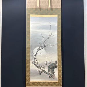 May include: A traditional Japanese scroll painting featuring a branch of a flowering tree with white blossoms against a soft, moonlit sky. The artwork is framed with decorative borders in shades of gold and green, and is suspended by a beige fabric.