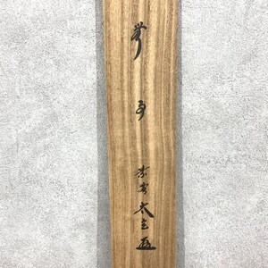May include: A wooden plank with visible wood grain and black Japanese calligraphy. The calligraphy is vertically aligned and includes several characters. The wood has a light brown color.