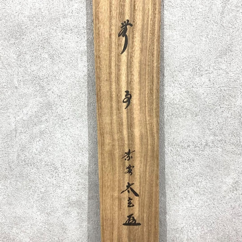 May include: A wooden plank with visible wood grain and black Japanese calligraphy. The calligraphy is vertically aligned and includes several characters. The wood has a light brown color.