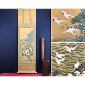 May include: A traditional Japanese scroll painting depicting white cranes in flight over a landscape with a blue tree and white flowers. The scroll has a gold patterned top and bottom.