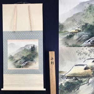 Könnte beinhalten: Japanische Rollbildmalerei, die eine neblige Berglandschaft mit einem kleinen Dorf zeigt. Das Kunstwerk weist gedämpfte Töne von Grün, Grau und Beige auf. Die Schriftrolle ist auf einem hellblauen Stoff mit einem gepunkteten Muster und rosa Akzenten befestigt.