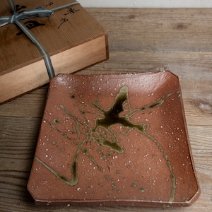 May include: A square, reddish-brown ceramic plate with a unique design. The plate features a dark green, abstract splatter pattern and small white speckles. A wooden box with a blue ribbon is in the background.