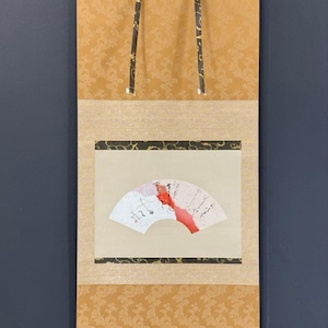 May include: A traditional Japanese scroll painting featuring a fan design with red and white accents. The scroll has a gold floral patterned background and black trim. The scroll is suspended by two black and gold hanging cords.