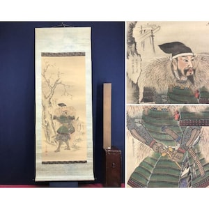 Op de afbeelding: Een antiek Japans rolschilderij dat een samoerai-krijger in een winterlandschap afbeeldt. Het kunstwerk heeft gedempte kleuren, met de krijger in groene bepantsering en een met bont gevoerd kledingstuk. Een houten kist en een houten stok staan naast de rol.