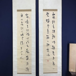 Op de afbeelding: Twee verticale Aziatische rolschilderijen met zwarte kalligrafie op crèmekleurig papier. De rollen hebben houten staven aan de boven- en onderkant. De achtergrond is donkerblauw.