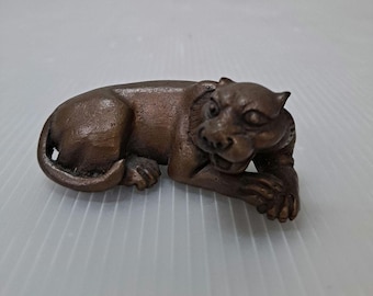 May include: A bronze-colored sculpture of a resting feline, possibly a panther or jaguar. The animal is depicted lying down with its front paws extended and head raised, showcasing detailed facial features and a curled tail.