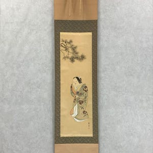 Op de afbeelding: Japans rolschilderij met een vrouw in kimono onder een dennentak. Het kunstwerk gebruikt gedempte kleuren, met de nadruk op de kleding en uitdrukking van de figuur. De rol is omlijst met een patroonrand en heeft een ophangkoord.