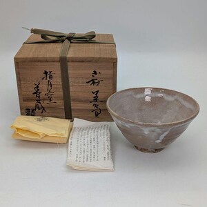 May include: A ceramic bowl with a light brown glaze and white streaks, presented with a wooden box tied with a green ribbon. The box has Japanese characters on it, and the set includes a yellow cloth and a paper with text.