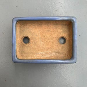 May include: A rectangular bonsai pot with a light blue rim and a natural brown interior. The pot has two drainage holes. The pot is designed for growing small trees and plants.